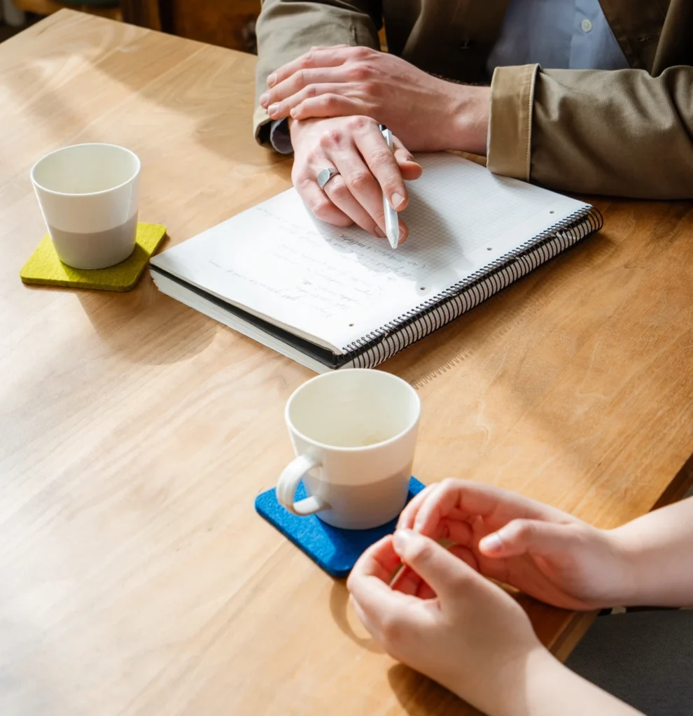 Twee professionals in gesprek aan een houten tafel met notitieboek en koffie, kenmerkend voor de begeleiding bij PEX Academy