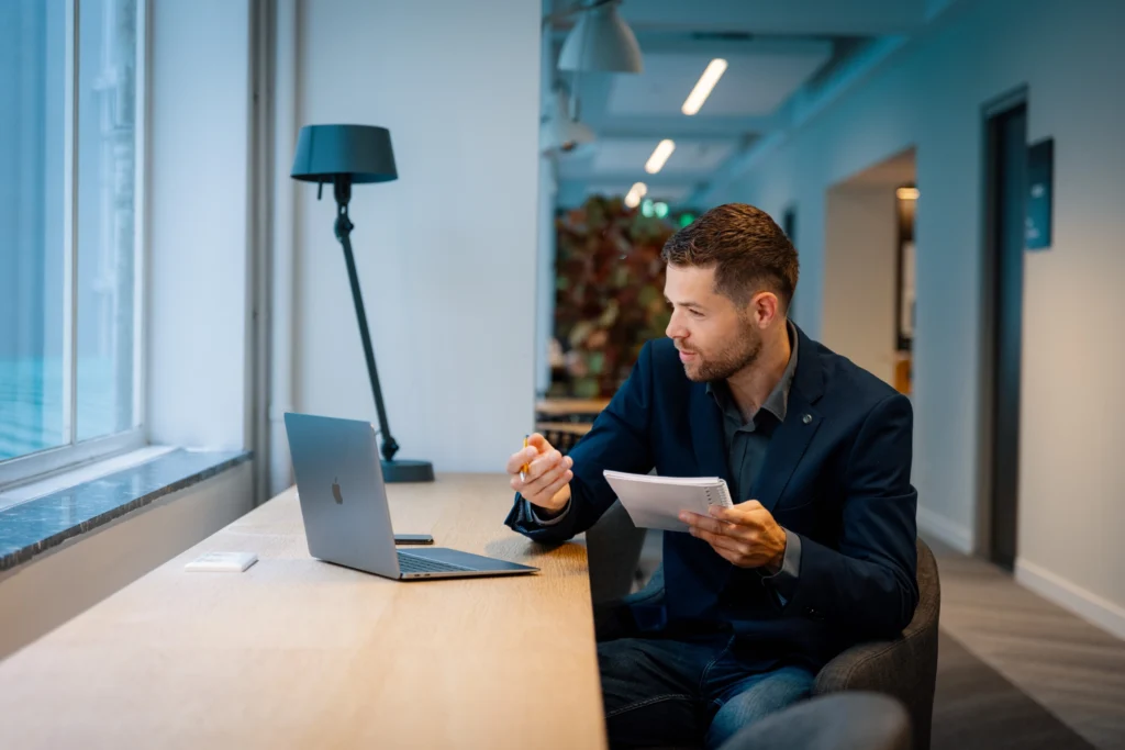 Man in zakelijk jasje maakt aantekeningen tijdens een opleiding sociaal domein achter een laptop in een modern kantoor